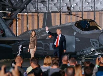 Trump, heavyset man in suit with red tie waving next to wife Melania, shapely woman with long brown hair standing in front of a military plane with people in foreground facing him