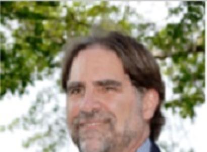 Middle aged white man with short brown hair, a grayish beard and mustache wearing a blue suit standing in front of a tree