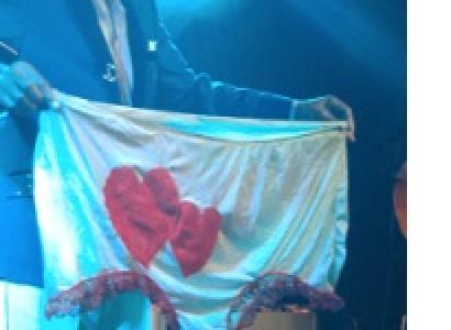 Smiling black man standing in a suit on a stage holding up a huge pair of women's undies that are white with two red harts on them