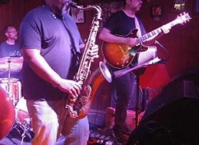 Heavy set black man in a cap playing the saxophone with other guys in a band onstage