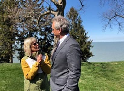 Short blonde wtoman wearing a yellow coat and sunglasses talking and gesturing to a taller white man with gray hair and a gray suit outside by tree and a lake 