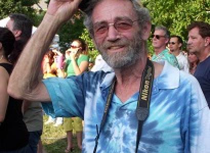 White man raising up his cap to the camera, smiling with sunglasses and gray mustache and beard, wearing a blue tye-dye t-shirt standing outside around trees and a lot of other people 