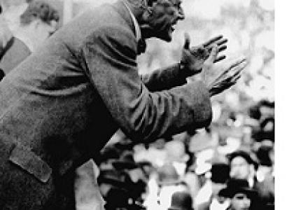Black & white photo of a thin bald man sideways speaking and gesturing with arms outstretched to a crowd of people