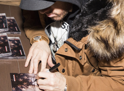 Guy in a black hoodie and brown heavy winter coat with fur collar sitting at a table with his face looking down where he is signing a CD