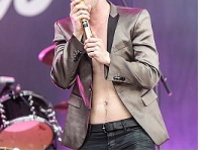 Young white man with satiny suit jacket and no shirt with shaggy brown hair singing intoa ic