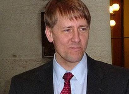 White man in a dark suit, white shirt, red tie with shaggy blonde hair with a pensive worried look