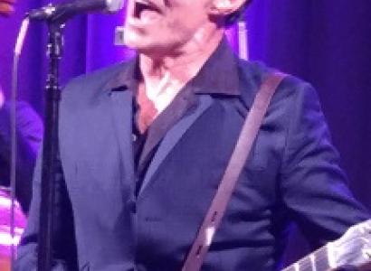 White man at a microphone belting out a song wearing a dark suit and shirt unbuttoned with a guitar strapped around him