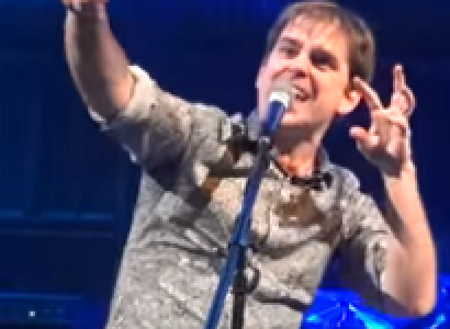 Young white man with both arms up in the air singing at a mic