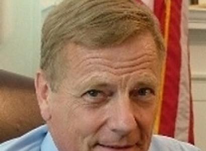 Close up of white man's face with brown hair and blue button down shirt and tie in front of a flag, his eyebrows a bit raised