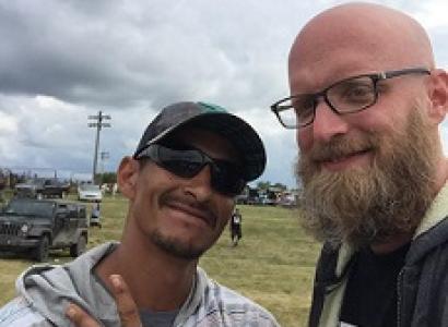 Bearded white man with glasses an bald head smiling with a dark-skinned man in sunglasses and a baseball cap outside in front of some cars and a cloudy sky