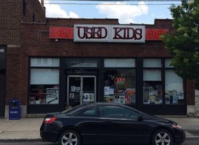 Brick building with sign at top saying Used Kids and a storefront with windows, a black car parked in front