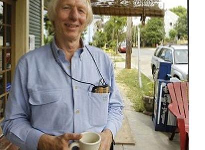 Thin tall white man with white hair smiling holding a coffee cup standing outside a store with a outdoor chair