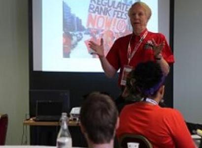 Gray haired man in red shirt waving his arms in front and talking to a group seated before him with a projected image on a screen behind him