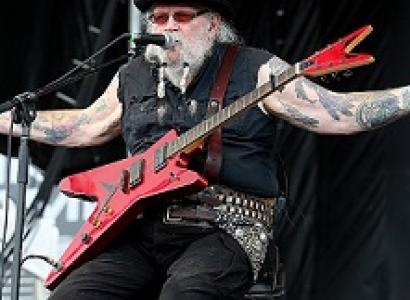 Older man with black hat, white hair and beard, sunglasses, sitting on a chair on stage with his arms spread wide with lots of tattoos and a red rock and roll electric guitar on his lap
