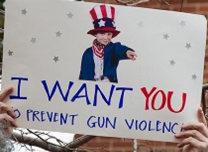 Someone holding up a sign with a picture of a young boy dressed like Uncle Sam and the words I WANT YOU to prevent gun violence
