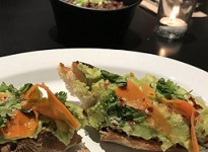Two pieces of hard bread with green and orange foodstuff on them on a white plate with a bowl and glass on the table in the background