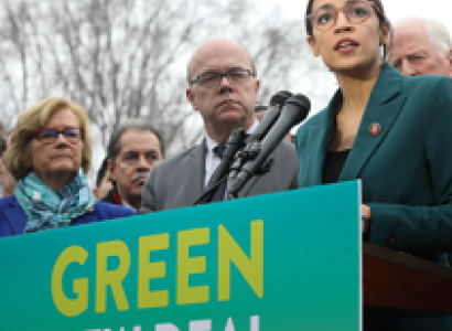 Sign saying Green New Deal with people behind it and young woman with glasses speaking
