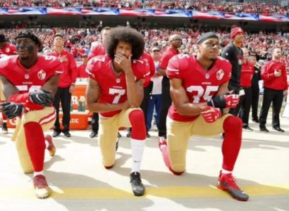 African American football players with red jerseys and tan pants kneeling on one knee in a stadium full of people