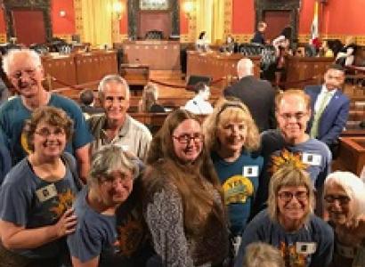 Lots of smiling white people male and female leaning toward each other all wearing blue T-shirts in a city council chambers