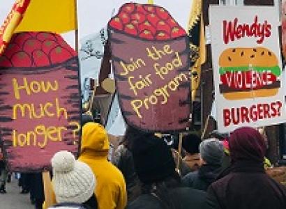 People marching with very large high signs with depictions of barrels of tomatoes with the words How much longer? and Join the Fair Food program and a Wendy's hamburger that says violence on it