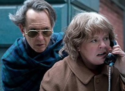 A man with sunglasses and hair blowing in the wind hovering behind a woman looking upset talking into a pay phone