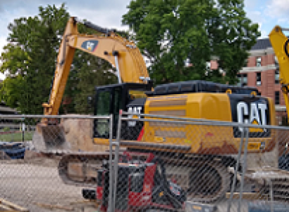 Big yellow machine truck with big scoop on a long neck that says CAT on the side behind a wire fence with other machinery and trees in the background