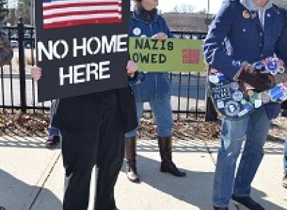 Someone holding a sign with a US flag on it on black background saying Hate has no home here, a guy playing a guitar that's covered with stickers with political sayings and a woman with red hair behind in the middle with a sign against Nazis