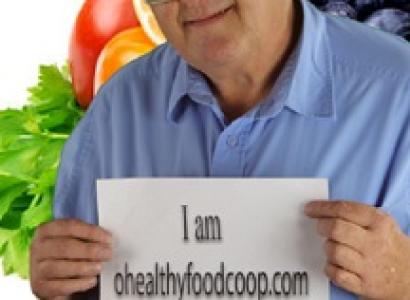 Older white man with glasses in front of fruits and vegetables holding a sign that reads I am healthyfoods.com
