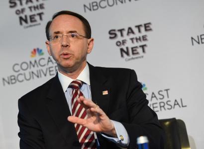 Balding white man with wire rimmed glasses wearing a suit speaking and make a hand gesture