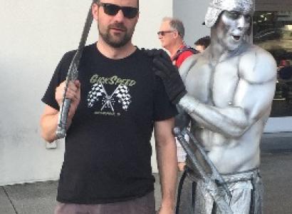 Guy with dark beard, sunglasses and sword next to a silver statue of a half-naked warrior with metal helmet
