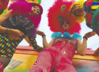 Camera angle looking from ground up at four decked out women with bright orange, pink and rainbow colored fluffy feathery bedazzled outfits