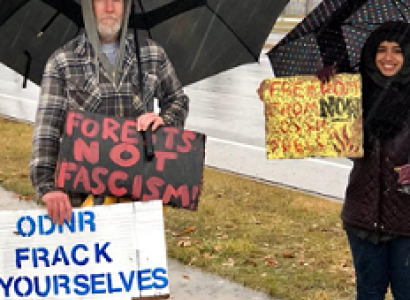 Two people standing outside in the rain holding umbrellas and signs, one says ODNR Frack Yourselves