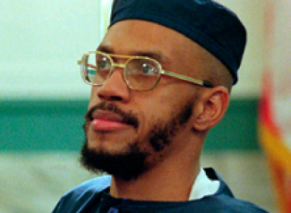 Black man with wire rimmed glasses and mustache and beard and a square topped blue hat