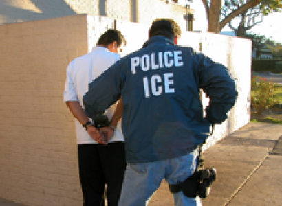 Back of man in blue jacket with white letters reading ICE putting another man with his back to us in handcuffs