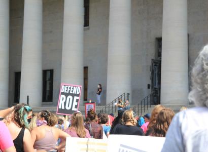 crowd protesting in front of state house with sign that says defend roe
