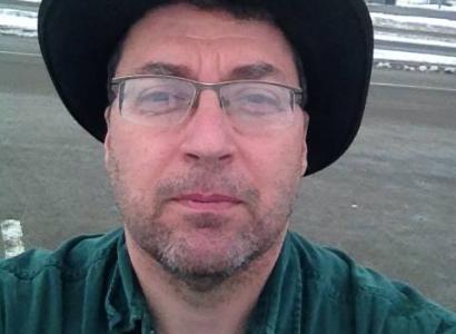 Man with glasses and wide brimmed black hat, looking grim