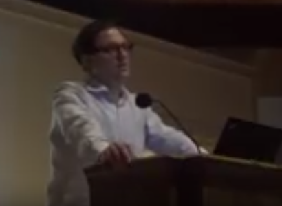 White man with glasses and white shirt standing and speaking at podium