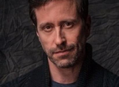 Thin faced man with brown hair and five o clock shadow looking quizzically at the camera