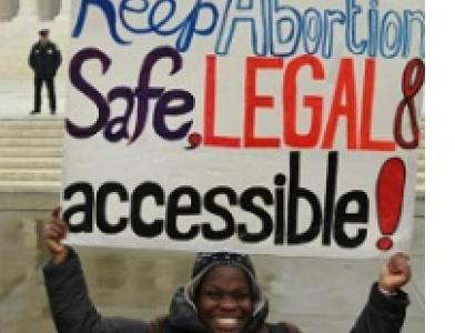 Young black woman smiling with winter clothes on outside an official looking government building holding a sign above her head saying Keep Abortion Safe, Legal & accessible! A cop is standing back in the distance.