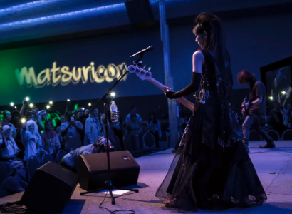 White woman in a Renaissance looking gown playing guitar on stage to a crowd standing under the sign Matsuricon