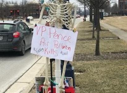 Skeleton leaning against a pole outside holding an anti-Pence sign