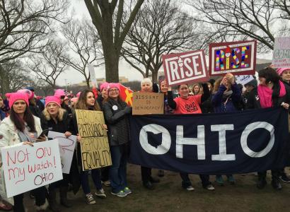 Women outside wearing pink winter hats holding signs and a big banner that says OHIO