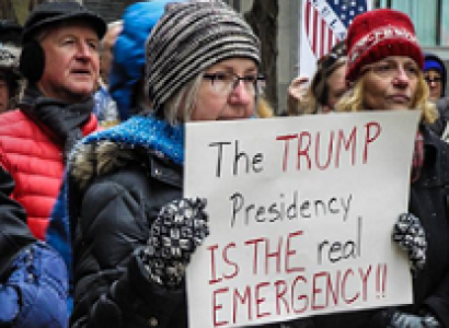 Woman in cold weather gear holding a sign at a protest outside that reads The Trump Presidency is the REAL emergency