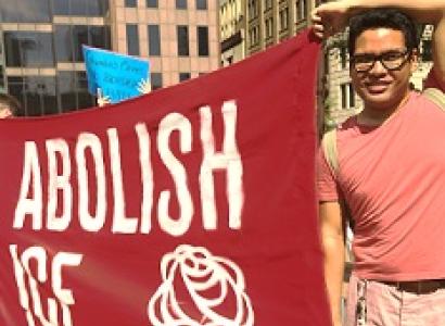 Young dark haired man with glasses holding a large red sign that says Abolish ICE with a DSA symbol of fist and rose
