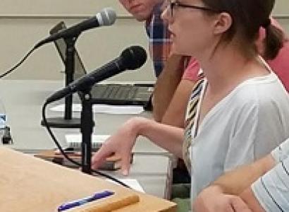 Young white woman with glasses and brown hair tied back speaking and gesturing at a podium with two guys at the table behind her 