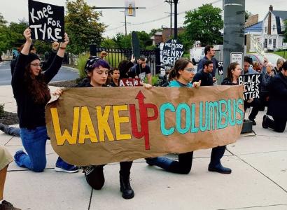 Young white people holding a sign saying Wake Up Columbus kneeling on ground and as part of a rally with lots of people behind them one with a sign saying Knee for Tyre