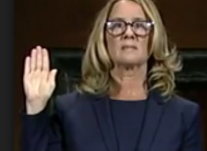 Blonde woman with glasses holding her hand up as if swearing to tell the truth