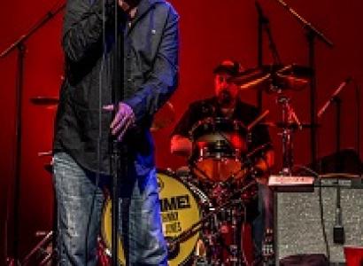 Middle aged white man with red sunglasses on singing with wide mouth open into a standing mic in front of a guy playing drums