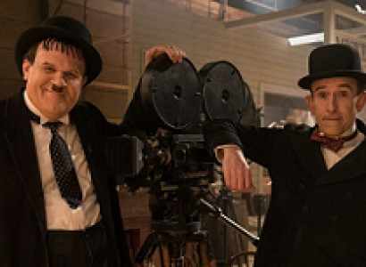 One large and one small man both in black suits with white shirts and hats leaning on an old time film projector