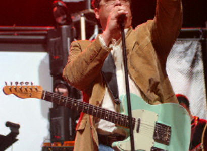White brwon-haired man with glasses singing at a mic with a guitar around his neck and he's pointing up in the air